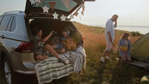 Kids Using Binoculars in Car Trunk while Parents Setting Up Camp on Lakeshore Stock Footage 304463088
