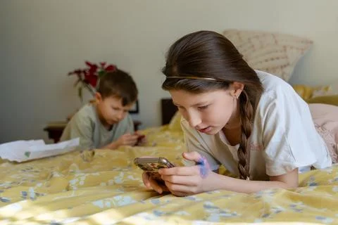 Kids using smartphones while lying on bed. A girl and a younger boy lie on a bed Stock Photos