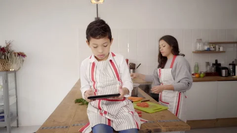 In the kitchen: child playing video games with a tablet while his mother cuts Stock Footage 150675953