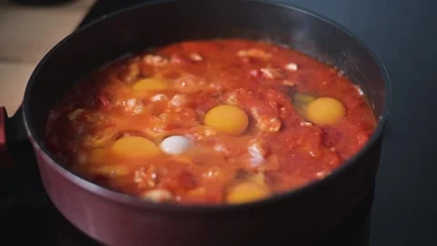 In a kitchen, eggs are being cooked in a bubbling tomato sauce in a frying pan. Stockbeeldmateriaal 344667523
