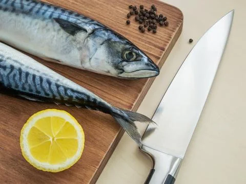 Kitchen knife, two raw mackerels, black peppercorns and lemon on cutting board Stock Photos