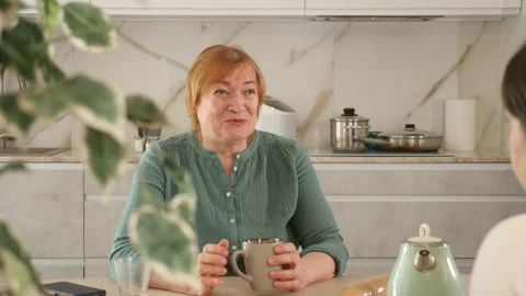 In kitchen at table, elderly smiling woman communicates with friend and drinks Vídeo Stock 303674301