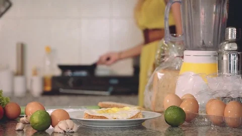 Kitchen table in foreground, woman making fried egg in background Video stock 77904276