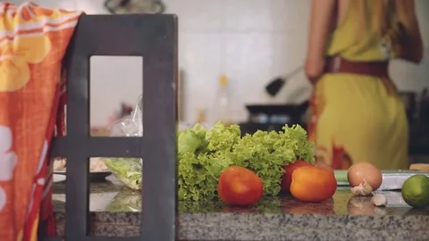 Kitchen table with lettuce and tomato, woman cooking on stove in background Stock Footage 77904239
