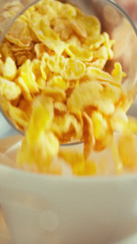 At a kitchen table a person pours crunchy cornflakes into a white bowl assem Stock Footage 331795192