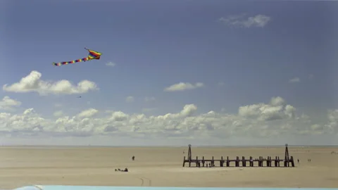 Kite hovering, people explore derilict pier on vast sandy English beach, 4K Stock Footage 238559097