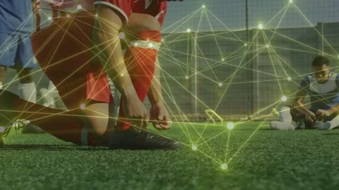 Kneeling red-kit player tying cleat on turf before play, node network tracking Stock Footage 333925009