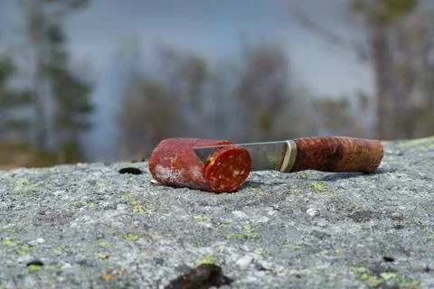 Knife with dry sausage on stone with blurry landscape background Stock Photos