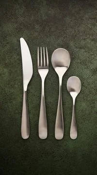 Knife, spoon, fork, teaspoon lying in a row on a dark green textured table Stock Photos