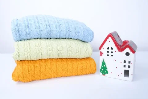 Knitted stacked piled sweaters, pullovers with winter ceramic house on white Stock Photos