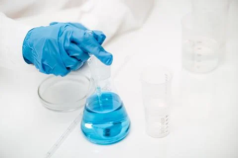 Lab assistant in rubber gloves pointing to flask with blue liquid on table 스톡 사진