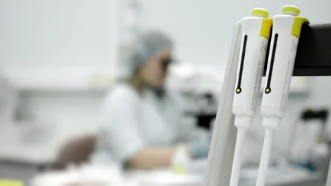 Laboratory micropipettes with technician working in background. 動画素材 321576044