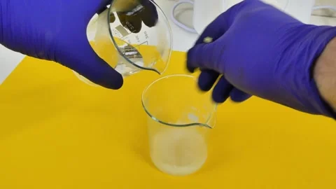 Laboratory technician in blue gloves pouring a liquid chemical into a beaker to Видео 334822100