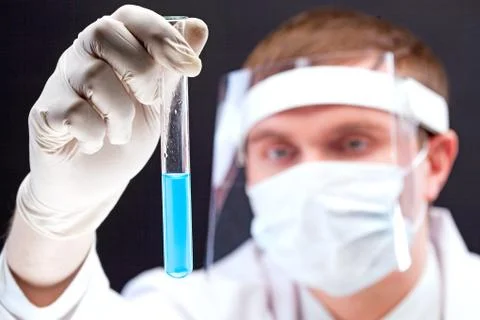 Laboratory worker looking at flask with blue liquid through transparent safe mas Stock Photos