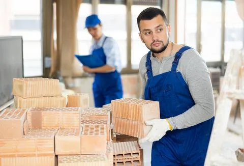 Laborer in blue overalls stacking bricks at construction site indoors Stockfoto's
