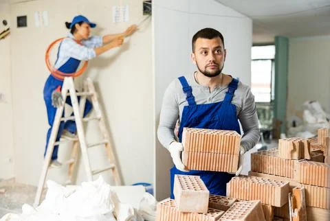 Laborer in blue overalls stacking bricks at construction site indoors Stockfoto's