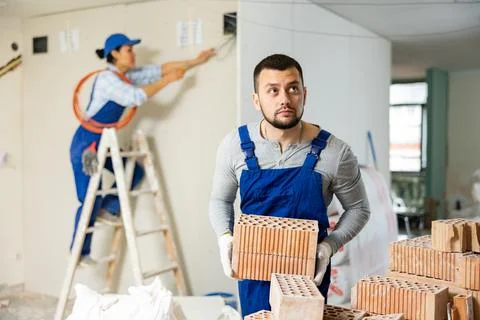 Laborer in blue overalls stacking bricks at construction site indoors Stock Photos