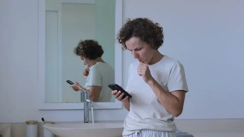 Lady brushing teeth in bathroom and scrolling on smartphone. Wide shot of woman Stock Footage 257752027