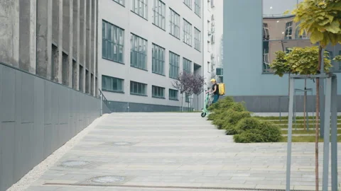 Lady delivery worker with backpack and yellow cap rides electric scooter along Stock Footage 171890814