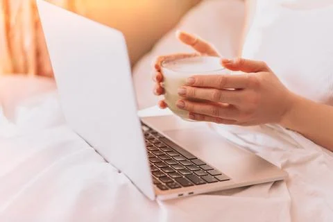 Lady drinking coffee while working on a laptop in a white bed Stock Photos