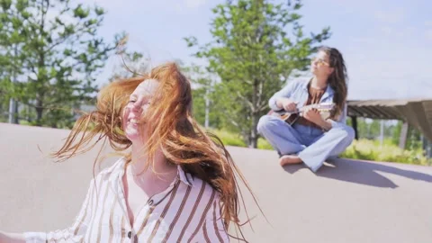 Lady jumps up with joy while friend plays ukulele in city garden Stock Footage 147578395