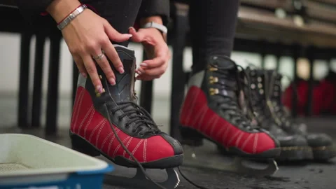 Lady putting on red black ice skates tightening laces on wet floor near bench Видео 321006983