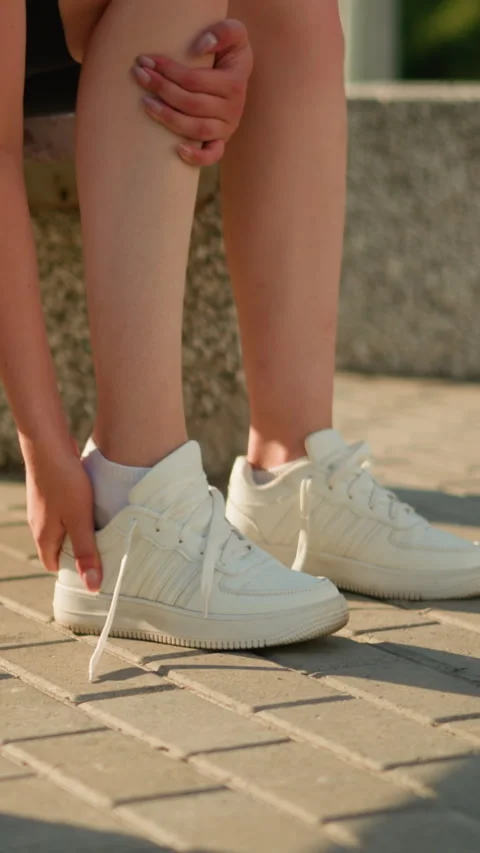Lady Removing Sneaker Seated on Bench Outdoors with Greenery in Background Stock Footage 310520985