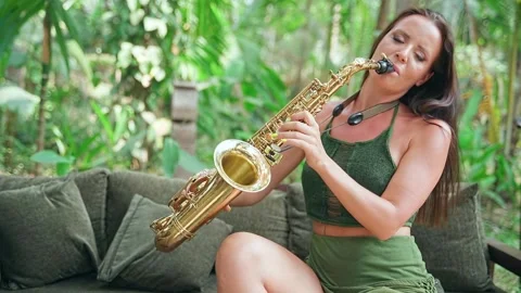 The lady sits on the couch in a long green dress and plays the gold saxophone. Video stock 234021960