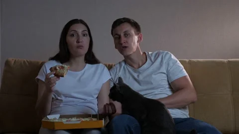 Lady sits by man on sofa eats pizza feeding husband and cat Stock Footage 163873634