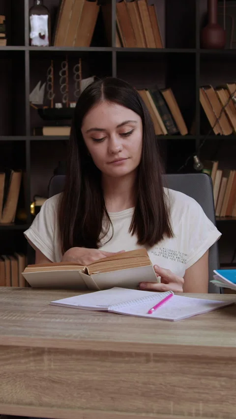 Lady student reads book at table sitting by huge bookshelf filled with Video stock 305408309