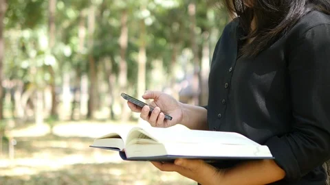 Lady using mobile phone and holding book over green garden background Video stock 101001632