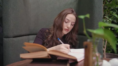 Lady Writing in Book While Glancing at Open Book on Table in Cozy Green Space Stock Footage 310698101