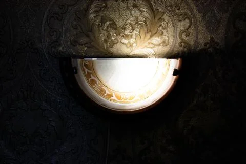 Lamp is on the wall. Lit in the half-light. Ornate wallpaper. Stock Photos