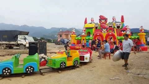 Lao child rides circular plastic train, kids on inflatable bouncer, man walks Видео 76926533