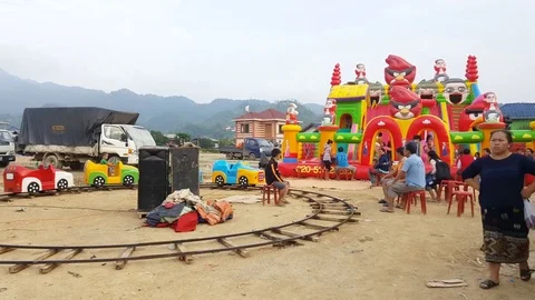 Lao child rides circular plastic train, kids on inflatable bouncer, woman walks 스톡 동영상 76926875