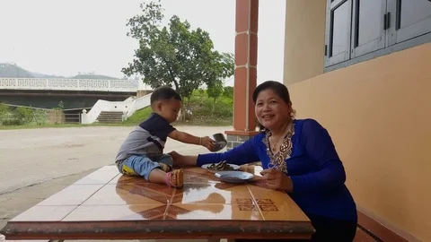 Lao infant sits on table, plays with bowl, grandma, bridge, trees in background Vídeos de archivo 76804809