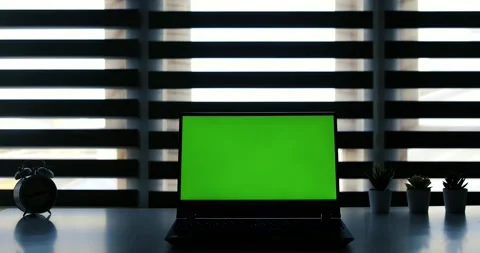 Laptop with green screen on white table in front of roller blind. Dark office 库存影片 148157217