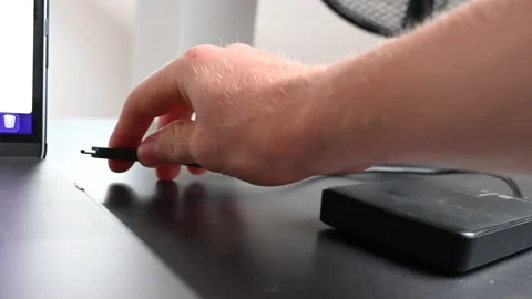 A laptop user plugs an external hardrive with a black cable to his Stock Footage 213178669