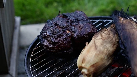 A large beef brisket and corn in the husk being barbequed on a small charcoal Stock Footage 111447242