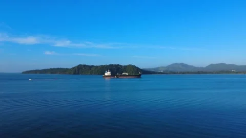 Large cargo ship sails across a calm blue sea towards lush green islands under a Stock Footage 325009854