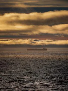 Large cargo ship sails across the ocean under dramatic golden clouds at sunse 스톡 사진