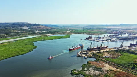 Large cargo ships navigate a river near a busy port with green land in the Stock Footage 341583351