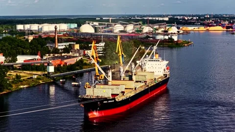 A large cargo vessel moves through a busy industrial port, surrounded by Stockbeeldmateriaal 325570400