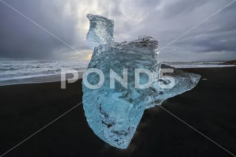Large chunk of ice sitting on the shore of Iceland with dramatic skies in behind Stock Photos