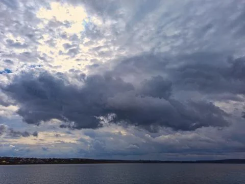 Large clouds float across the blue sky low above the river and the village Stock Photos
