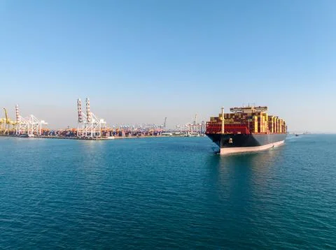 Large Container Cargo Ship Navigating Near Industrial Shipping Terminal At .. Foto stock