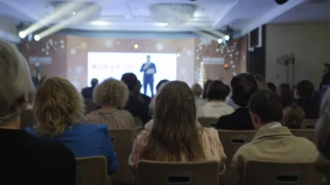 Large crowd of people are sitting in chairs in a room, watching a speaker Stock Footage 301941049