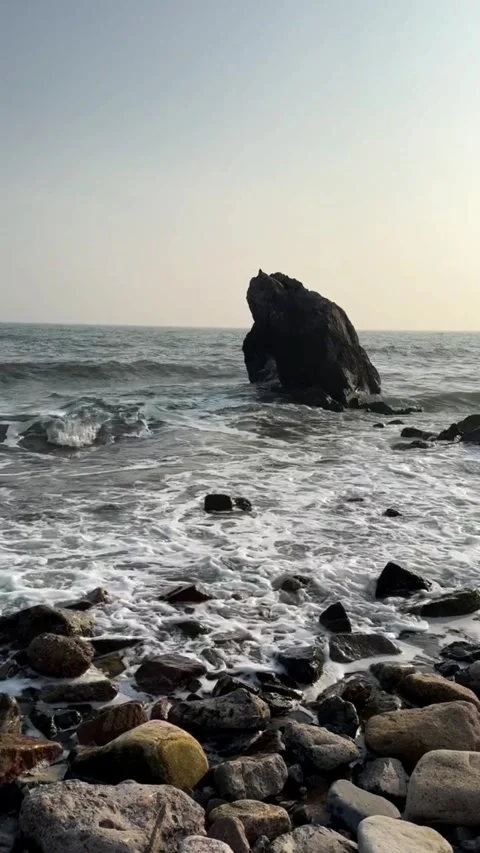 A large dark rock formation standing in the ocean waves on a rocky beach Vidéo 334163435