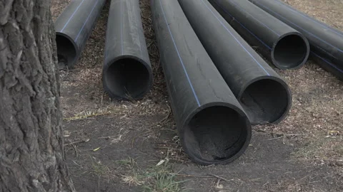Large diameter plastic black pipes lie on the ground outside near a tree Stock Footage 286027665