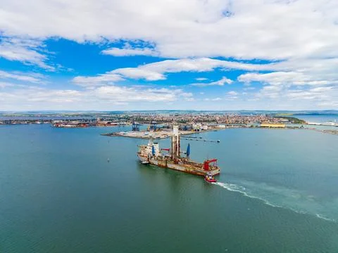 Large drilling rig moves through calm waters near a busy port with clouds i.. 写真素材